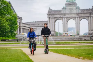 Two persons, one on a Dott bike, the other on a Bolt kick scooter, riding in front of the Cinquantenaire Arch